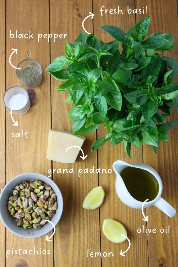 image of ingredients for pesto with pistachios: fresh basil leaves, a bowl of pistachios, grana padano cheese, organic olive oil and a lemon